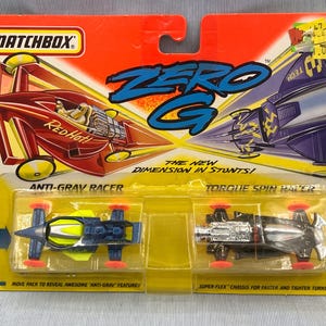 May include: Matchbox Zero G toy car set featuring a red and black Anti-Grav Racer and a black and silver Torque Spin Racer. The packaging features the text "The New Dimension in Stunts!" and "Move pack to reveal awesome "Anti-Grav" features!"
