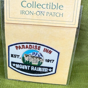 May include: Collectible iron-on patch featuring the Paradise Inn at Mount Rainier. The patch has a blue border and includes the text "Paradise Inn", "Mount Rainier", and "Est. 1917". The patch is on a tan background.