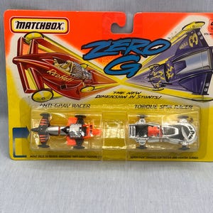 May include: Matchbox Zero G toy set featuring two race cars, "Red Hot" and "Torque Spin Racer", on a yellow and orange card. The card reads "The New Dimension in Stunts!" and "Anti-Grav Racer".