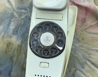 May include: A vintage white Trimline telephone with a rotary dial. The phone features a speaker, a handset, and a dial with numbers and letters. The phone is a classic design from the past.