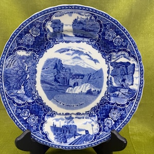 May include: A blue and white decorative plate featuring scenic views of Colorado Springs. The plate has a scalloped edge and is supported by a black stand. Text on the plate includes "Balanced Rock" and "Gateway of the Garden of the Gods."