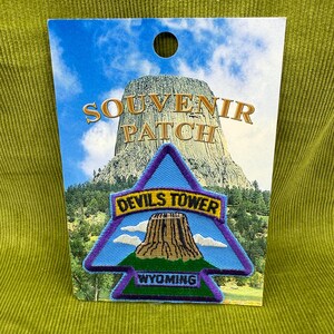 May include: A souvenir patch featuring Devils Tower in Wyoming. The patch is triangular with a purple border and a blue background. The text "Devils Tower" and "Wyoming" are displayed on the patch. The card reads "Souvenir Patch".