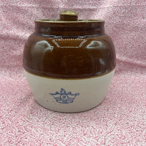 May include: A two-tone ceramic jar with a lid. The upper portion is brown, and the lower portion is off-white. A blue crown design with the number "2" is on the front. The jar is likely for storage.