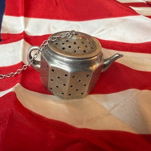 May include: A silver-toned metal tea infuser shaped like a miniature teapot with a hexagonal body and a perforated surface. The infuser features a small handle, a spout, and a lid, all attached to a thin metal chain with a circular ring at the end.