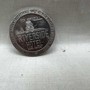 May include: Silver-colored casino token with the text "One Dollar Gaming Token - Acceptable Only at Pick Hobson's Riverside Reno, Nevada" and an image of a car driving on a road.