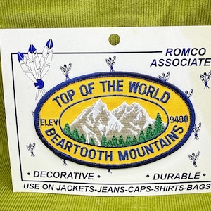 May include: A patch with the text "TOP OF THE WORLD BEARTOOTH MOUNTAINS" in yellow and blue, featuring a mountain scene. The patch is on a white card with text that says "DECORATIVE" and "DURABLE".