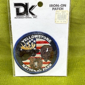 May include: An iron-on patch featuring a circular design with the text "YELLOWSTONE NATIONAL PARK." The patch includes illustrations of a moose, bison, and bear, set against a backdrop of the American flag. The patch is on a white backing.