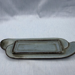 May include: A handmade, ceramic serving tray with a rectangular inner dish and a larger, uniquely shaped outer tray. The tray is a muted blue-gray color with dark brown trim, perfect for serving appetizers or small dishes.