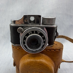 May include: A vintage black and silver camera with a leather case. The camera has a lens with the word "HIT" and "MADE IN JAPAN" on it. The leather case is brown with a strap.