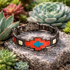 May include: A bracelet with a Southwestern design. It features a central red and turquoise cross-like shape, with white and red squares. The metal has a dark, antique finish. The bracelet is made of metal.