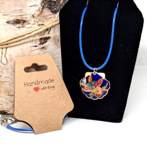 Puede incluir: Collar con cordón azul y colgante con diseño floral en tonos rojos, rosas y dorados, sobre fondo azul oscuro. El colgante se exhibe en un soporte negro, con una etiqueta marrón que dice "Handmade with love".