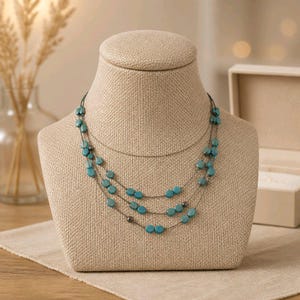 May include: A three-strand necklace with turquoise-colored beads. The necklace is displayed on a beige jewelry bust. The beads are oval-shaped and strung on a dark metal chain. The necklace is a fashion accessory.