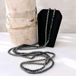 Op de afbeelding: Een lange, meerstrengige ketting bestaande uit kleine, donker metallic gekleurde kralen met een gefacetteerde afwerking. De ketting is gedrapeerd in een los, cirkelvormig patroon, wat de reflecterende kwaliteit van de kralen en de consistente, uniforme grootte van elke individuele kraal over de gehele lengte van de streng laat zien.