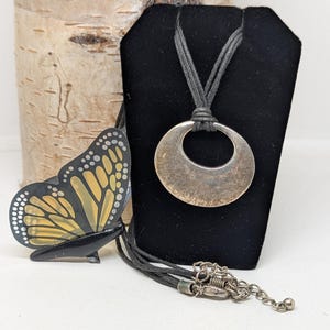 Pode incluir: Um colar com pingente prateado com um design de círculo aberto grande. O colar pende de um cordão preto. À esquerda do colar, há um objeto decorativo em forma de borboleta preta e amarela.