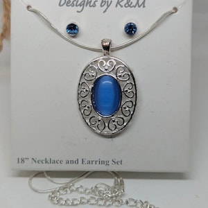 May include: An 18-inch necklace and earring set. The set includes a silver-toned chain necklace with a pendant featuring a blue oval stone within a decorative silver filigree design. Matching blue stud earrings are also included. The packaging reads "Designs by K&M".