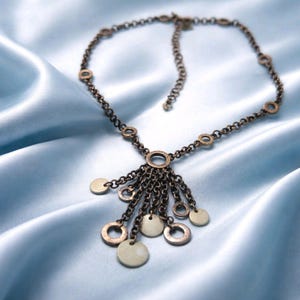 May include: A bronze-coloured necklace with a chain and pendant design. The necklace features small, round, gold-coloured accents and a series of dangling chains with circular and disc-shaped pendants. The jewellery is displayed on a light blue satin fabric.