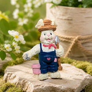 May include: A white rabbit figurine wearing a brown straw hat, a red neckerchief, and blue overalls with a pink heart detail. The rabbit holds a silver garden trowel in one hand and carries a small pink bucket on its other arm, standing upright.