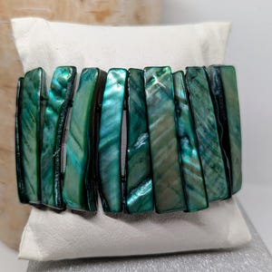 Puede incluir: Un brazalete verde iridiscente con segmentos rectangulares pulidos. El brazalete se exhibe sobre una almohada blanca, mostrando los tonos verde mar brillantes del material. El diseño del brazalete es simple, pero elegante.
