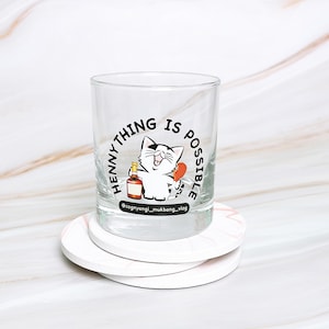 May include: A clear glass tumbler with a black and white illustration of a cat holding a bottle of liquor. The text "Henny Thing Is Possible" is printed around the cat. The tumbler is sitting on a white coaster.