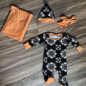 May include: A baby clothing set featuring a black and white patterned footed onesie with orange trim, a matching hat, a headband, and a solid orange blanket. The set is designed for newborns.
