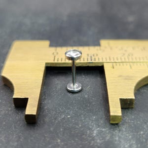 May include: A small, silver-colored labret stud with a flat, round top and base. The stud is positioned between the jaws of a brass caliper, suggesting a measurement. The background is a dark, textured surface.