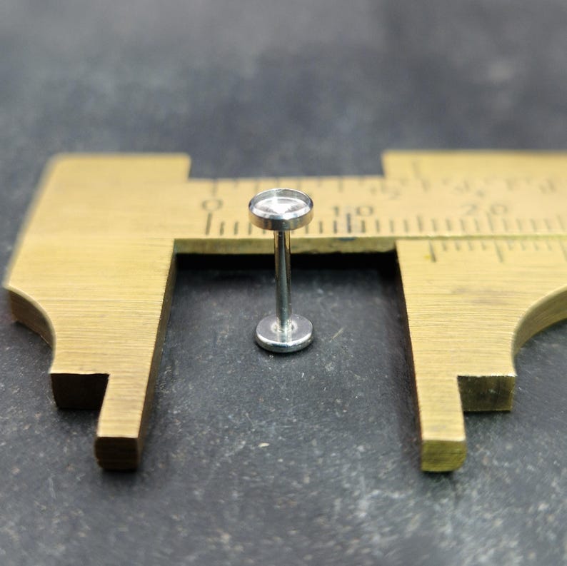 May include: A small, silver-colored labret stud with a flat, round top and base. The stud is positioned between the jaws of a brass caliper, suggesting a measurement. The background is a dark, textured surface.