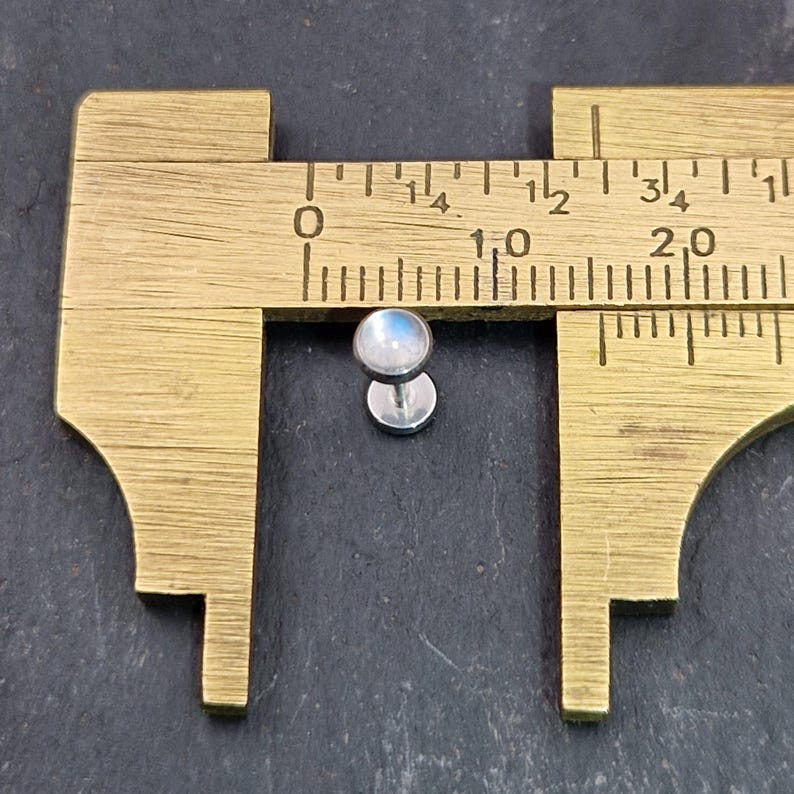 May include: A small, silver-colored stud earring with a round, iridescent moonstone cabochon. The earring is positioned next to a brass caliper, which measures the earring's size. The earring is approximately 0.25 inches in diameter.