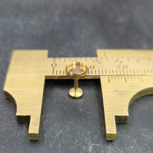 Könnte beinhalten: Ein kleines, goldfarbenes Miniatur-Cocktailglas aus Metall mit einer runden Schale und einem schlanken Stiel. Das Glas befindet sich neben einem Messschieber aus Messing, der zum Messen verwendet wird. Der Hintergrund ist eine dunkle, strukturierte Oberfläche.