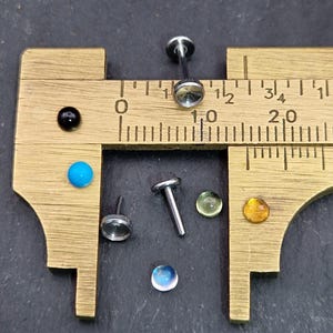 May include: A collection of body jewelry, including labret studs and gem-set tops, displayed with a brass caliper. The caliper shows measurements in inches. The jewelry features various colors, including clear, blue, green, and amber gems, and a black and turquoise bead.