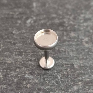 May include: A small, silver-colored jewelry setting with a round, shallow cup and a short, straight post. The setting is designed to hold a small stone or embellishment. The piece is shown against a dark, textured background.