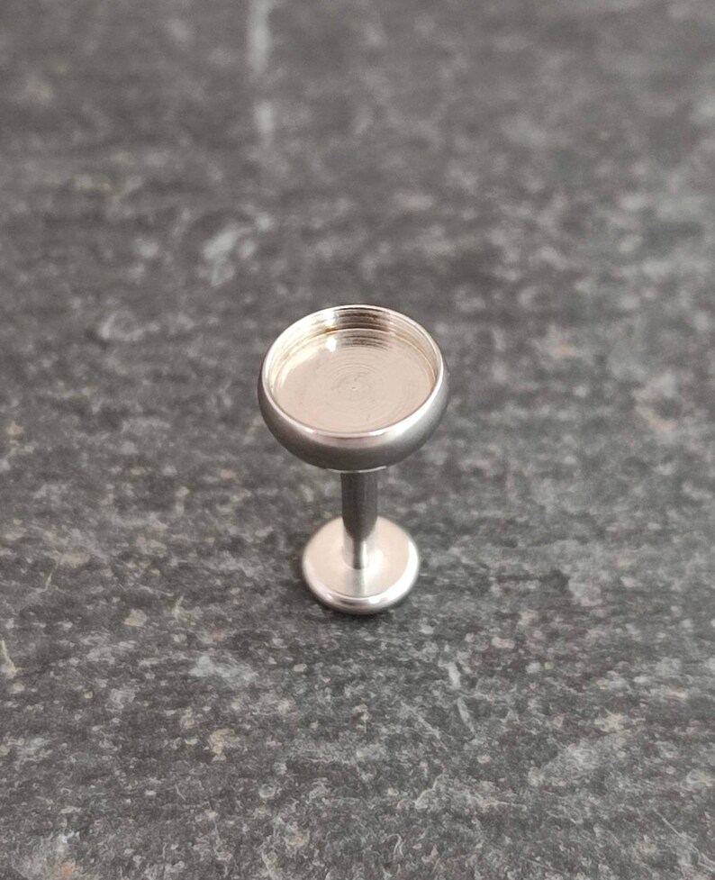 May include: A small, silver-colored jewelry setting with a round, shallow cup and a short, straight post. The setting is designed to hold a small stone or embellishment. The piece is shown against a dark, textured background.