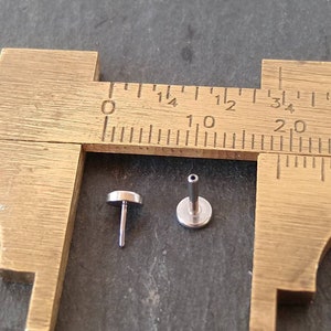 May include: Two small, silver-colored, flat-topped jewelry studs are displayed next to a brass caliper. The studs have a post and a flat, circular top. The caliper is marked with inch measurements.