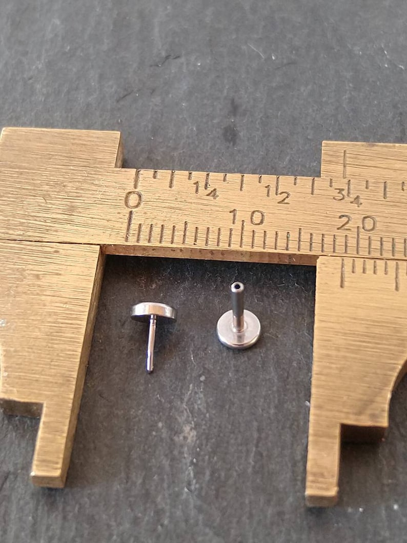 May include: Two small, silver-colored, flat-topped jewelry studs are displayed next to a brass caliper. The studs have a post and a flat, circular top. The caliper is marked with inch measurements.