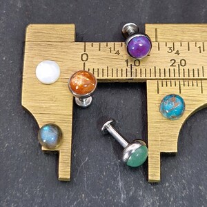 May include: A collection of gemstone stud earrings with various colors, including purple, orange, blue, and green. The earrings are displayed on a brass measuring tool, showcasing their size and detail. The studs have a silver-colored post.