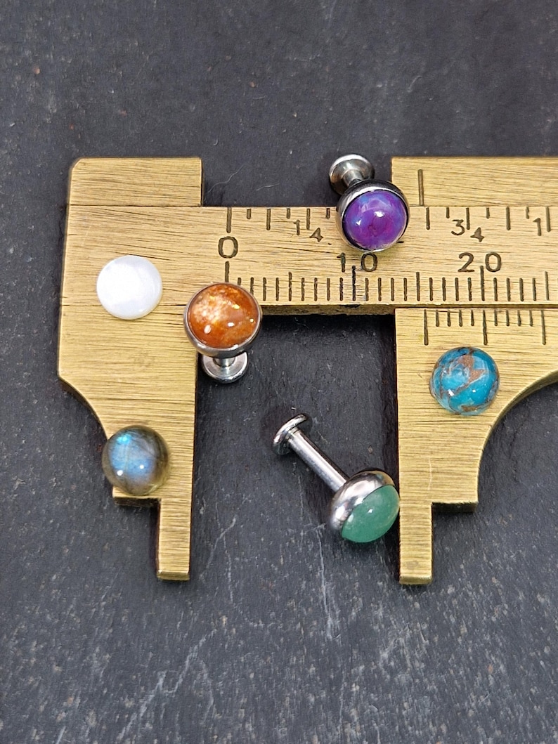 May include: A collection of gemstone stud earrings with various colors, including purple, orange, blue, and green. The earrings are displayed on a brass measuring tool, showcasing their size and detail. The studs have a silver-colored post.