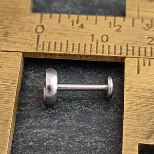 May include: A silver-colored labret stud with a flat, circular front and back, connected by a straight post. The jewelry is shown on a dark gray surface next to a wooden ruler.