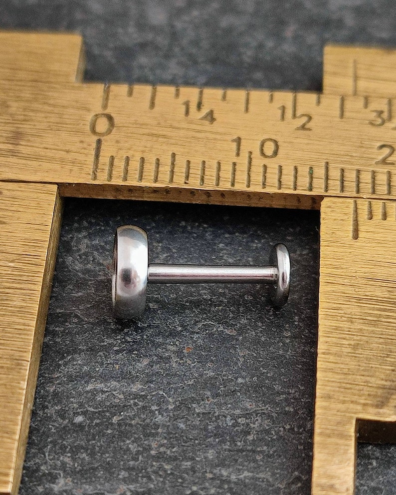 May include: A silver-colored labret stud with a flat, circular front and back, connected by a straight post. The jewelry is shown on a dark gray surface next to a wooden ruler.