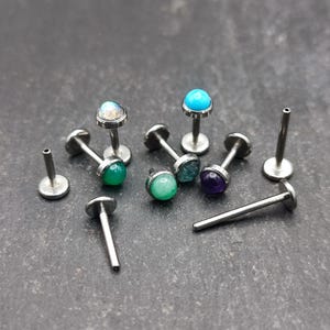 May include: A collection of small, silver-colored body jewelry pieces with colorful gemstone tops. The gemstones are various colors, including green, blue, and purple. Some pieces are complete, while others are posts.