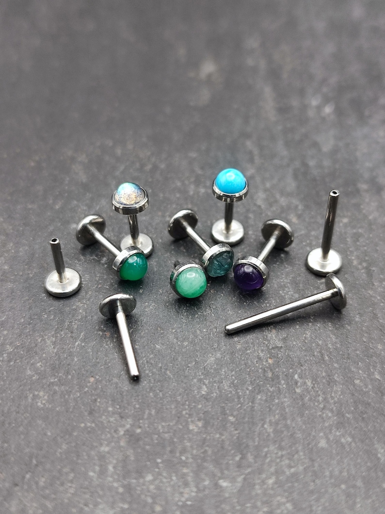 May include: A collection of small, silver-colored body jewelry pieces with colorful gemstone tops. The gemstones are various colors, including green, blue, and purple. Some pieces are complete, while others are posts.