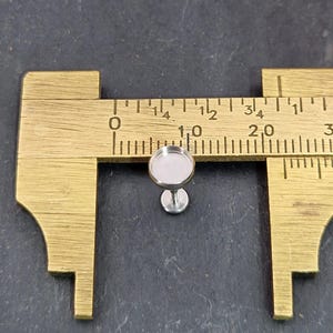 May include: A small, silver-coloured earring stud with a round bezel, shown next to a brass ruler. The stud's bezel measures approximately 1 cm in diameter. The earring post is attached to a small, circular base.