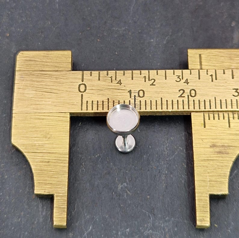 May include: A small, silver-colored earring stud with a round bezel, shown next to a brass ruler. The stud's bezel measures approximately 0.4 inches in diameter. The earring post is attached to a small, circular base.