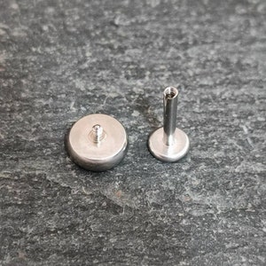 May include: Two silver-colored, flat-backed body jewelry pieces. One piece has a rounded, flat base with a threaded post. The other piece has a flat, circular base with a threaded post and a long, cylindrical post.