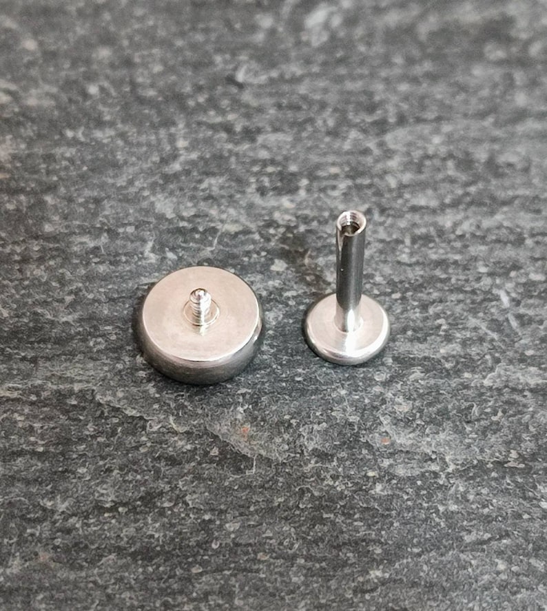 May include: Two silver-colored, flat-backed body jewelry pieces. One piece has a rounded, flat base with a threaded post. The other piece has a flat, circular base with a threaded post and a long, cylindrical post.