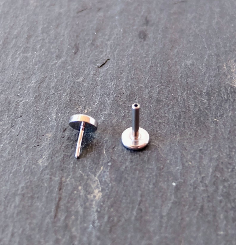 May include: Two silver-colored lip piercing studs. Each stud has a flat, circular top and a straight post. One stud is upright, showing the post and the underside of the top. The other stud is angled, displaying the top and post.