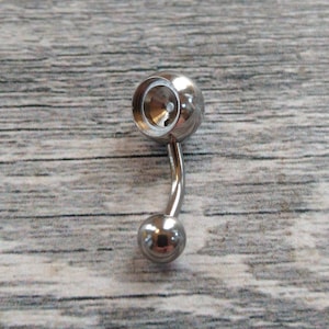 May include: A silver belly button ring with a round ball on one end and a larger ball with a circular opening on the other end.