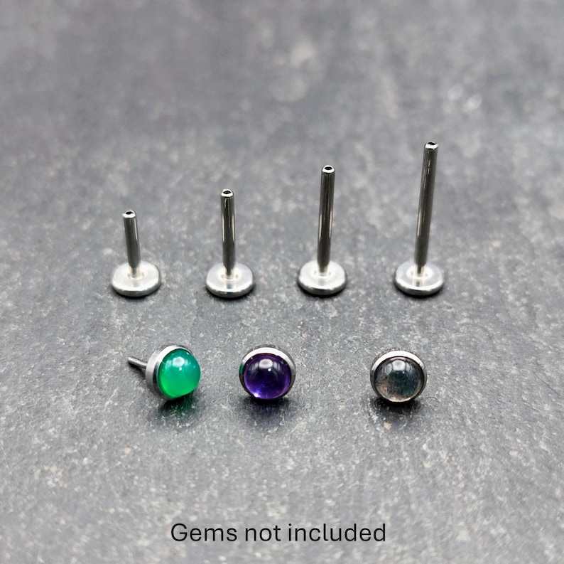 May include: A collection of silver-colored labret studs and posts, with three gemstone studs in green, purple, and gray. The text "Gems not included" is at the bottom of the image.