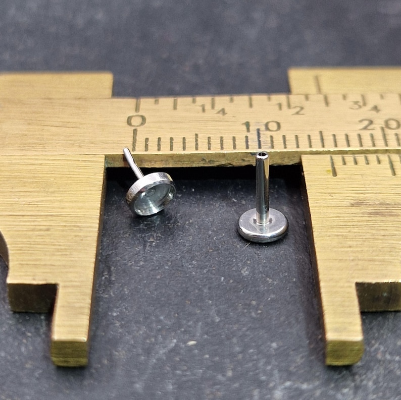 May include: Two small, silver-colored flat-back studs with a bezel setting, displayed on a brass ruler. The studs are approximately 0.25 inches in diameter. The background is a dark gray surface.