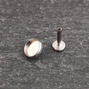 May include: Two pieces of body jewelry. One is a small, silver-colored, rounded bezel cup. The other is a silver-colored, flat-backed post with a threaded top. The jewelry is designed for body piercings.