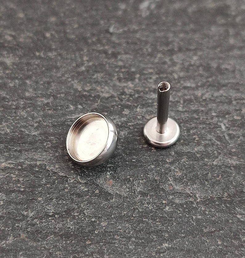 May include: Two pieces of body jewelry. One is a small, silver-colored, rounded bezel cup. The other is a silver-colored, flat-backed post with a threaded top. The jewelry is designed for body piercings.