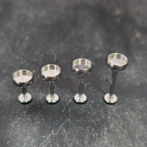 May include: Four silver-colored jewelry posts with a flat, circular top and a long, slender stem. The posts are arranged in a row, with the stems and tops of each post visible. The posts are on a dark, textured surface.